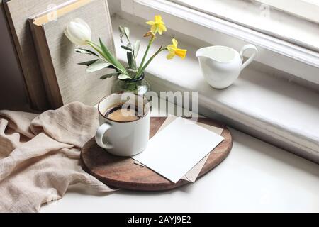 Cozy Easter spring still life. Greeting card mockup scene. Cup of coffee, books, wooden cutting board, milk pitcher and vase of flowers on windowsill. Stock Photo