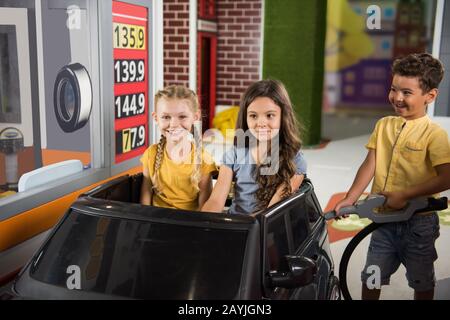 Little girls sitting in car at gas station in playroom. Stock Photo