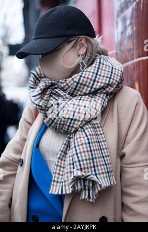 London, UK - 10 February, 2020 People wear mask for protection. Street style. Stylish woman in black dress, coat and a cap wears a beige mask Stock Photo
