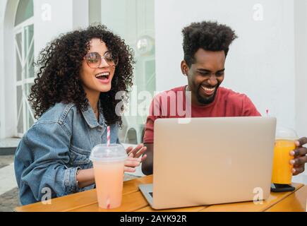 Portrait of two Afro friends using laptop while drinking fresh fruit juice outdoors at a cafeteria. Stock Photo