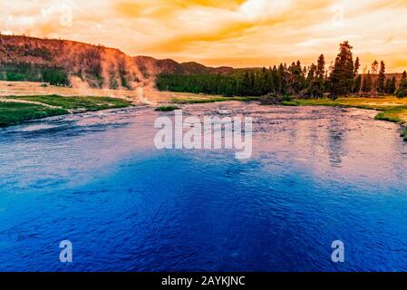 Deep blue lake with green meadows and forest under bright yellow sunrise. Stock Photo