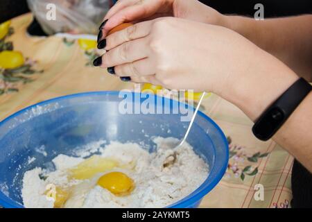 Woman with black manicure cooking and breaking eggs into a blue bowl of flour to make dough. Stock Photo