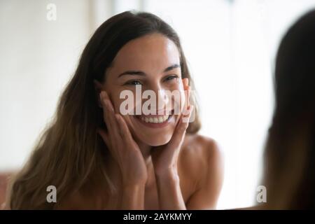 Smiling young woman look in mirror washing face Stock Photo