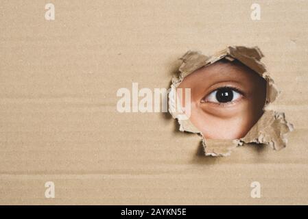 Boy eye peeking through a hole on a cardboard box with shocking gesture. Concept of mystery, secret and stalker Stock Photo
