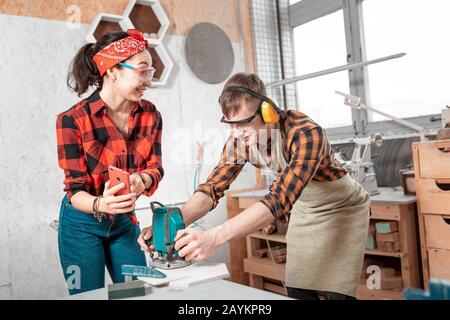 The woman customer showing smartphone to the carpenter who works with wooden products using an electric jigsaw. The concept of blue collar job Stock Photo