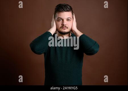 Young man with brown hair dressed casually in a green blouse standing over brown isolated background showing symbols of three wise monkeys hear no evi Stock Photo