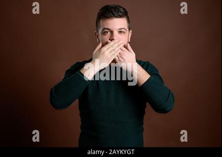 Young man with brown hair in green blouse standing over brown isolated brown background showing symbols of three wise monkeys speack no evil Stock Photo