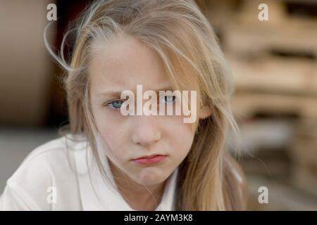 A sad, fair-skinned young girl with blond hair and blue eyes, pouting. The girl is whereing a white shirt. The image has a blurred background. Stock Photo