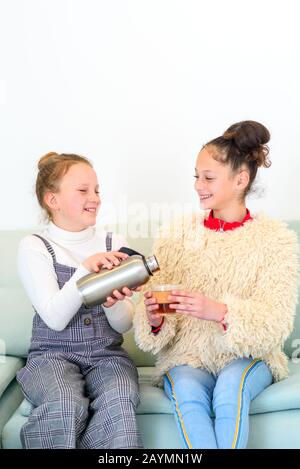 Health conscious teenager with reusable water bottle or tumbler tea pouring tea in reusable cup.Two teen with hot drink in new home.Young girls talking about funny things gossip laughing in apartment Stock Photo
