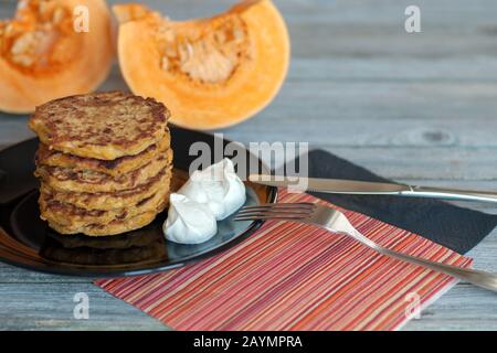Pumpkin pancakes stack on black plate, yougurt on the side, fork, knife, black and red napkins on wooden table, squash slices on background Stock Photo