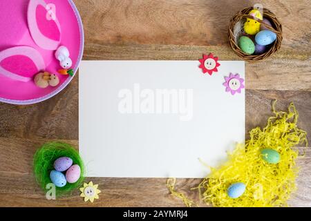 Colorful easter background flat lay on wooden table with white paper space for words. Pink hat, rabbit, chick, easter eggs in nest border. Design Stock Photo