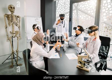 Five multiracial scientists or doctors using futuristic VR goggles headset and touching virtual screens, sitting at the table with microscope, tubes Stock Photo