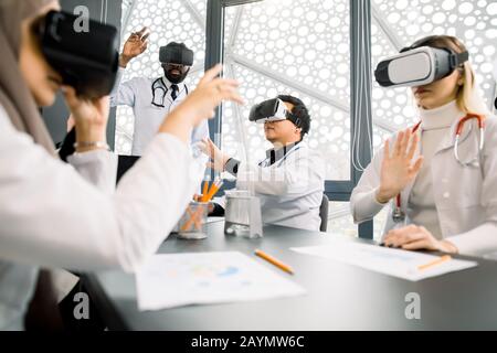 Team of multiethnical doctors, lab workers, scientists, Cacasian and Muslim women, African and Asian men, using virtual reality glasses for research Stock Photo