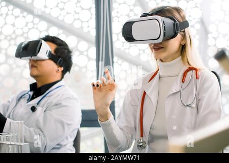 Young Caucasian woman and Korean man, doctors or scientists with stethoscopes using VR virtual reality glasses simulators, working with virtual Stock Photo