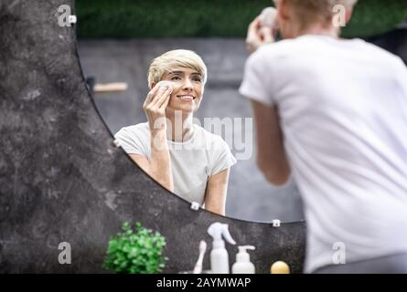 Lady Applying Skincare Product Using Cotton Pad Standing In Bathroom Stock Photo