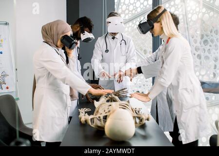 Working atmosphere in modern lab. Concentrated multiethnic professional biologists, doctors, scientists wearing VR glasses working with human skeleton Stock Photo