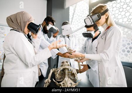 Group of young multiethnic medical students or doctors wearing VR glasses studying human skeleton anatomy in modern classroom. Medicine, anatomy Stock Photo