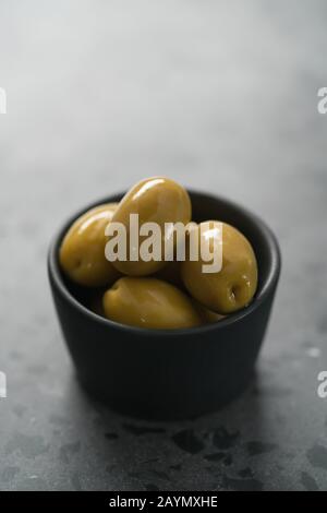 Whole big green olives in small black bowl on concrete background, shallow focus Stock Photo