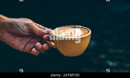 Hand Hold Cup Hot Latte Art in yellow mug against dark background.