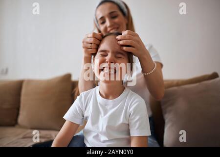 mother plaiting one braid for her child, woman doing kid's hgair, woman tightening kid's hair after the night. close up photo, funny girl making faces Stock Photo