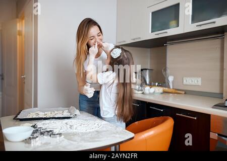crazy little adorable child is standing and touches her mothers nose with finger. smearing it with flour, close up photo, fun, entertainment, happines Stock Photo