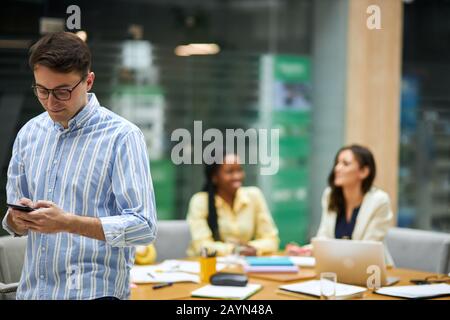 serious guy using his smart phone while his attractive female colleagues talking in the blurred background, gadget. close up photo, man sending e-mail Stock Photo