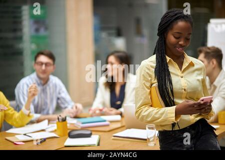 smiling positive girl in stylish yellow shirt chatting with friend, leaning on the office desk. blurred background, office workers sitting at the tabl Stock Photo