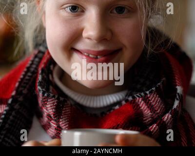 portrait of a sick child in a scarf and plaid with a cup of hot tea on the sofa in the apartment Stock Photo