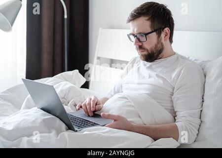 Freelancer morning. Focused man in pajamas works with a laptop, lying in a soft bed, in comfortable apartments, in natural light. Stock Photo