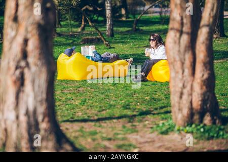 couple laying on yellow inflatable mattress in city park. reading book. drinking smoothies Stock Photo