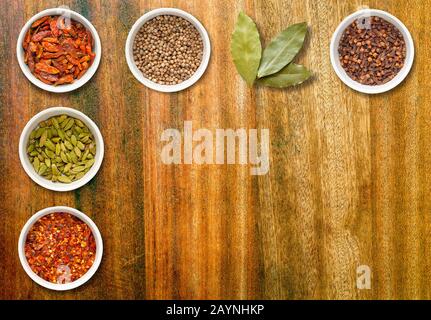 Spices including chilli flakes, cloves, bird's eye chillies, cardamoms, bay leaves and coriander seeds in white pots on an old dark wood chopping boar Stock Photo