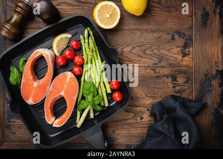 Uncooked salmon steaks and vegetables asparagus, tomato, lemon and basil on a grill pan, wooden table background with copy space. Healthy cooking. Top Stock Photo