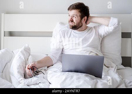 Creative process. Blogger distracted from working with a laptop, took off glasses and looks thoughtfully out the window in bed. Stock Photo
