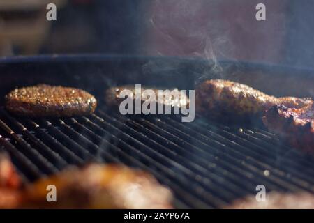 A portrait of the smoke in the middle of a hot barbecue grill. With the meat resting at the side of the bbq. There are hamburgers, chicken drumsticks Stock Photo