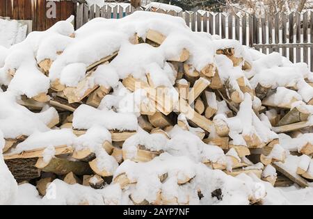 a pile, a stack of birch wood, logs, split with an axe, lie under the fresh fallen snow in the territory of a private village house near the fence Stock Photo