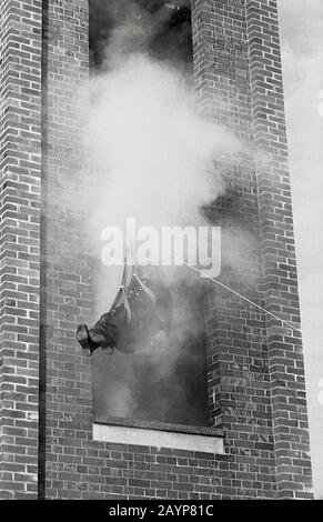 1960s, historical, a fireman taking part in a training exercise, sitting in a hoist, a pulley and rope, being evacuated from a smoking drill tower, Fleetwood, England, UK. Stock Photo