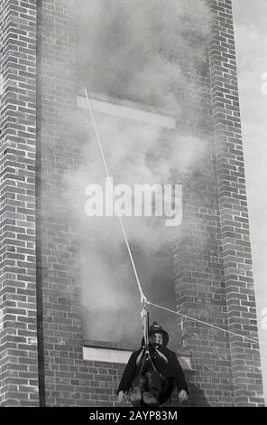 1960s, historical, a fireman taking part in a training exercise, sitting in a hoist, a pulley and rope, being evacuated from a smoking drill tower, Fleetwood, England, UK. Stock Photo