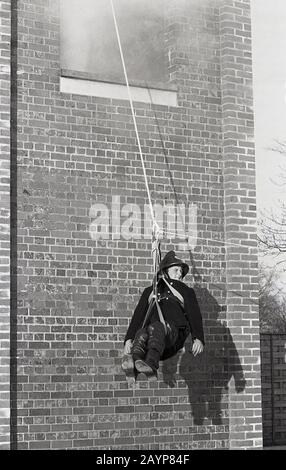 1960s, historical, a fireman in the uniform and helmet of the era, taking part in a training exercise in the grounds of the fire station, sitting in a hoist, a pulley and rope, being evacuated from a smoking drill tower, Fleetwood, England, UK. Stock Photo