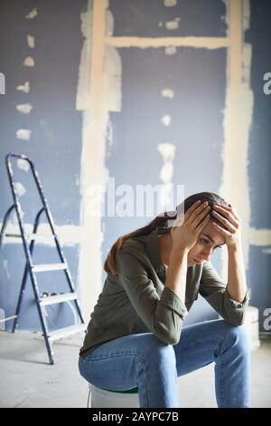 Vertical portrait of frustrated young woman devastated by renovations project sitting on paint can against dry wall, copy space Stock Photo