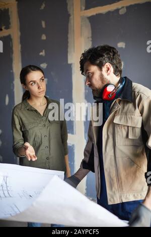 Portrait of young woman discussing floor plans with building contractor while standing against dry wall in under construction house, copy space Stock Photo