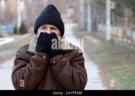 Elderly man standing on the rural street and warms up by covering his face with his mittened hands. Concept of cold weather, flu virus, frosty winter Stock Photo