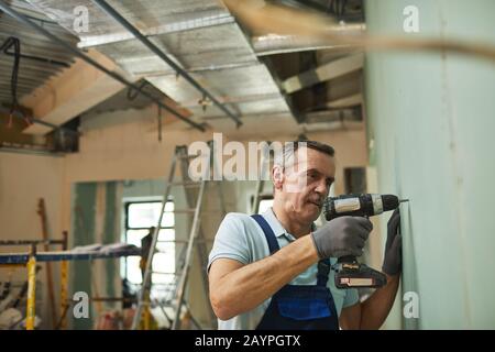 Side view portrait of senior construction worker drilling wall while renovating house alone, copy space Stock Photo