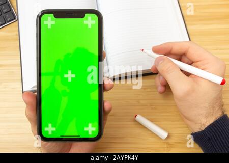 Mockup image of hand holding black mobile phone with green screen and red liner on the office table on the background with stationery tools. San Stock Photo