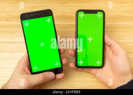 Mockup image of hands holding two black mobile phone with green screen on the office table on the background. San Francisco, February 2020 Stock Photo