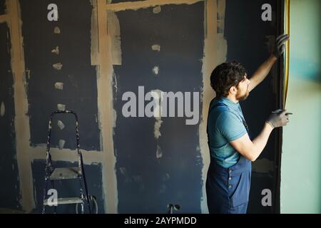 Side view of bearded construction worker measuring wall with tape while building or renovating house, copy space Stock Photo