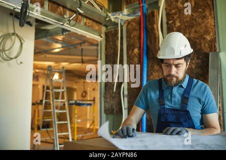 Waist up portrait of bearded construction worker wearing hardhat and looking at floor plans while renovating house, copy space Stock Photo