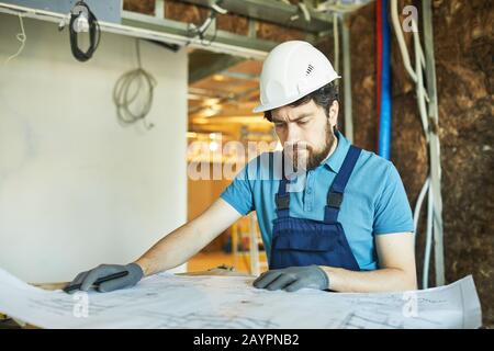 Portrait of bearded construction worker wearing hardhat and looking at floor plans while renovating house, copy space Stock Photo
