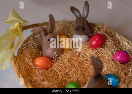 Colored easter eggs and caps of brown felt bunnies in nest of straw with colorful sweet sugar eggs. Felt bunnies as warm holder for easter eggs. Close Stock Photo