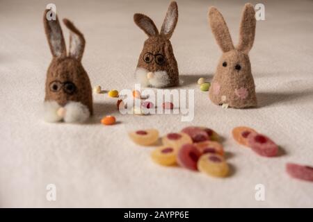 Three brown felt bunnies are sitting between colorful candy eggs on white ground. focus in background. Stock Photo