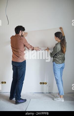 Full length back view portrait of married couple holding wallpaper to wall while redecorating house, copy space Stock Photo